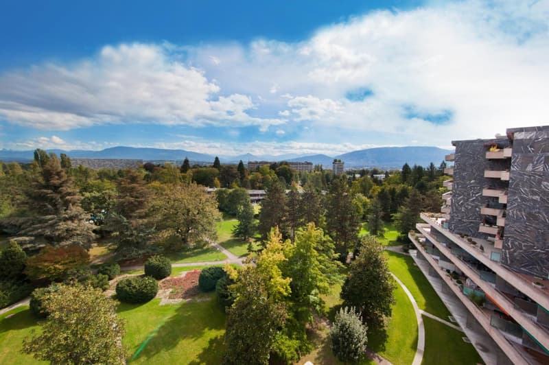 Multi-story residential building surrounded by greenery, with distant mountain range in the background.