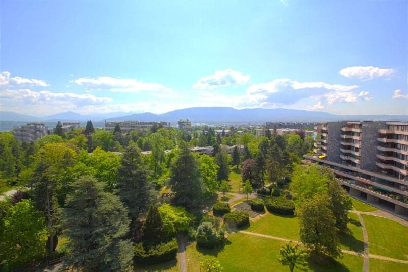 aerial view of a garden with lawn, trees, and buildings with mountains in the background