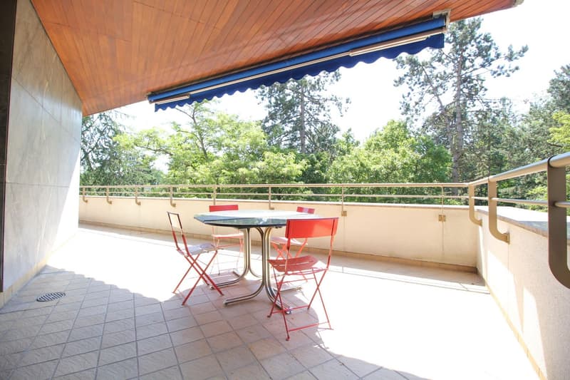 A balcony with a tiled floor, wooden ceiling with an awning, and a metal railing. There is a round table with four red chairs on the balcony. The balcony offers a view of the trees outside.