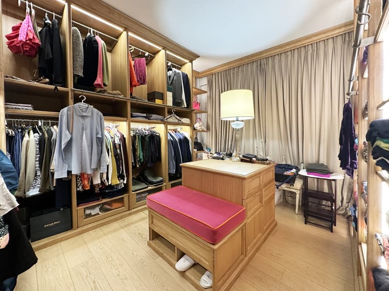 large closet filled with clothes, wooden bench, wooden dresser, pink cushion, white lamps, wooden floor