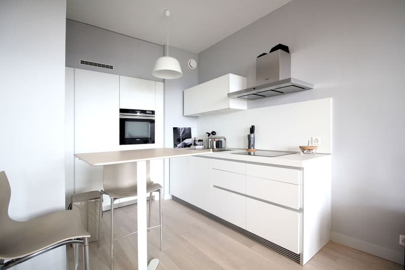 modern kitchen with white cabinets, wooden floor, island with stools, built-in oven, dishwasher, and a range hood