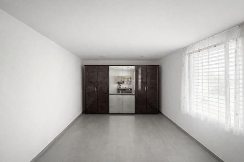 A room with gray flooring, white walls, and a large wardrobe with mirrored doors. It includes a built-in mirror on the left side, a window with white curtains, and a modern style.