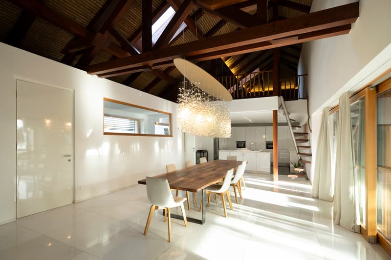 Dining room with wooden table, white chairs, white walls, wooden ceiling, chandelier, staircase, kitchen, window, mirror