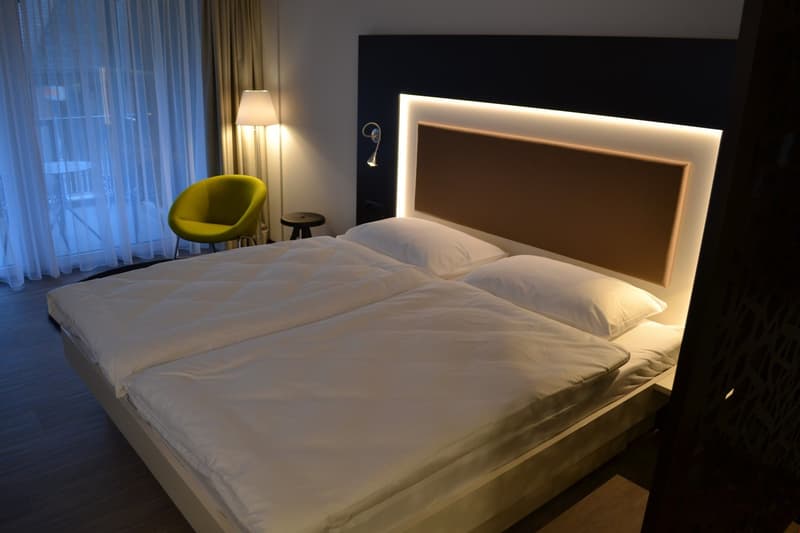 Spacious bedroom with a large bed, white bedding, and a modern, backlit headboard. There is a yellow armchair and a small side table with a lamp. The room has large windows with curtains, suggesting a balcony or terrace area.