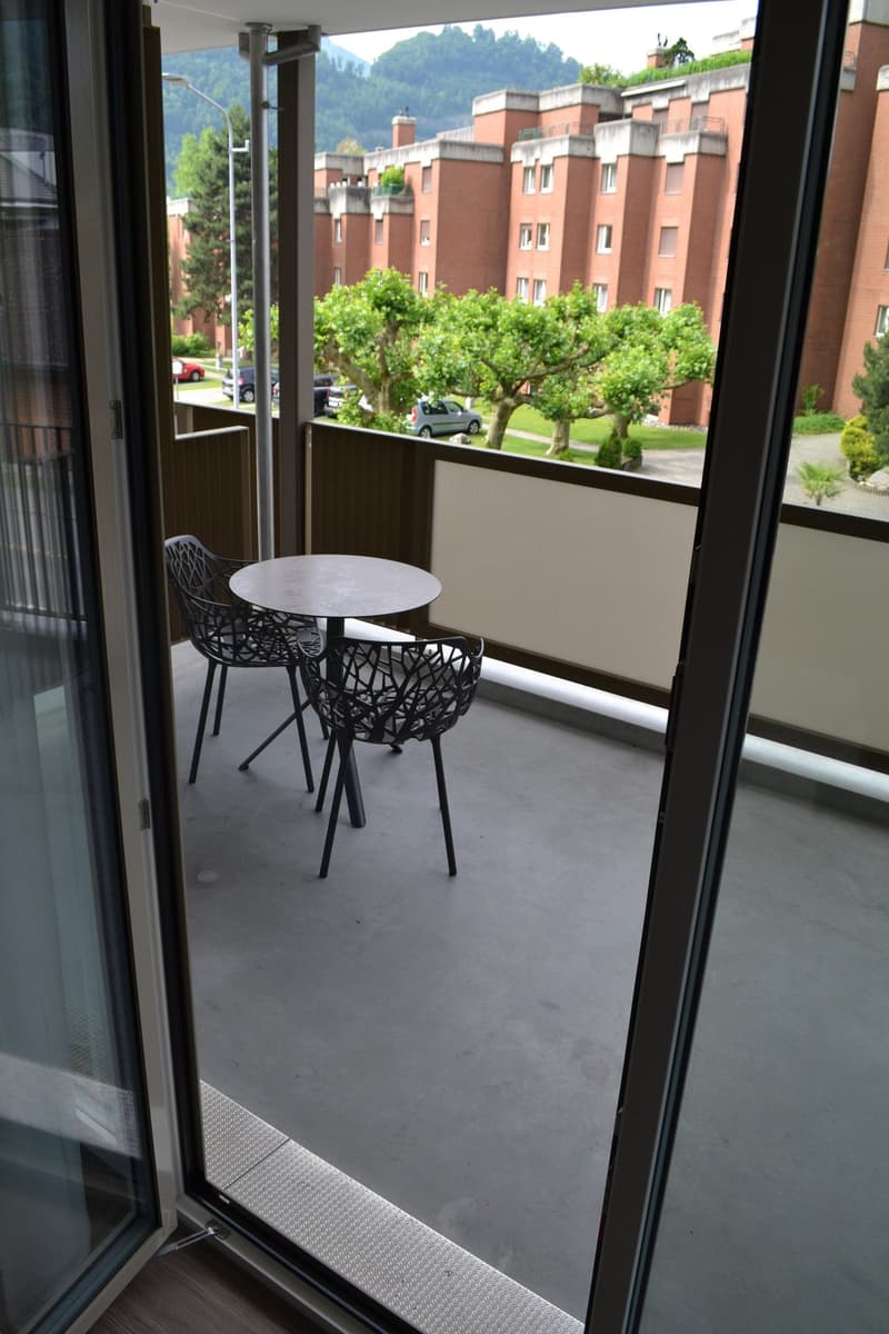 Balcony with a small table and two chairs, overlooking a residential area with multi-story brick buildings and trees.