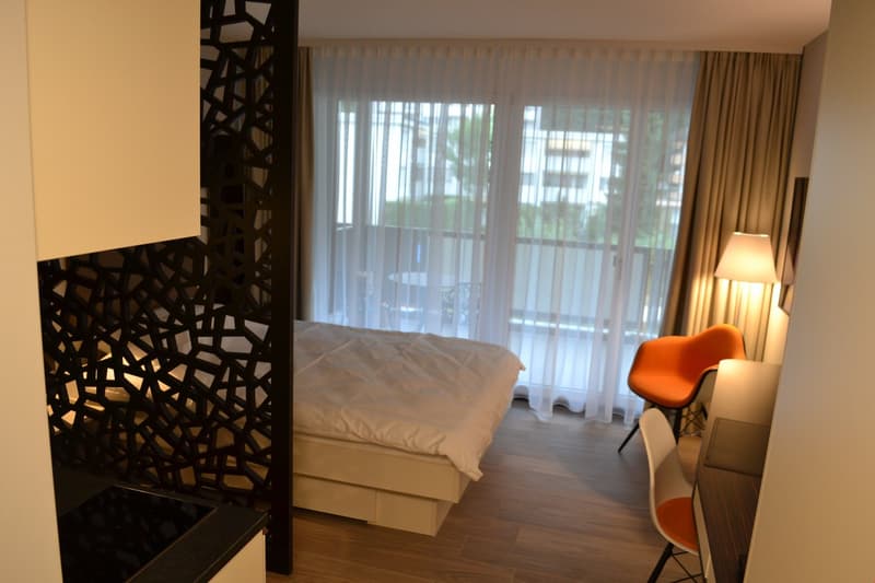 Spacious bedroom with large windows, wooden floor, white bedding, and an orange accent chair. The room features a decorative black partition wall and has access to a balcony or terrace.