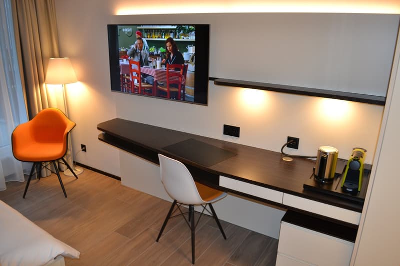 Modern living room with a large TV mounted on the wall, a dark wooden TV stand with drawers, an orange accent chair, and a white chair at a desk area. The room has hardwood flooring and appears to have a balcony or terrace visible through the window.