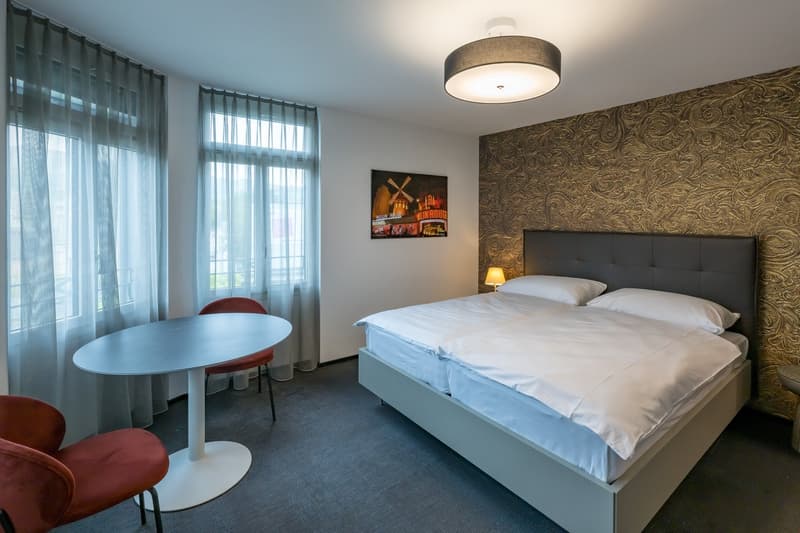 Spacious bedroom with large windows, modern furnishings including a bed with a headboard, a round table with chairs, and a patterned accent wall. The room has a cozy and stylish atmosphere.