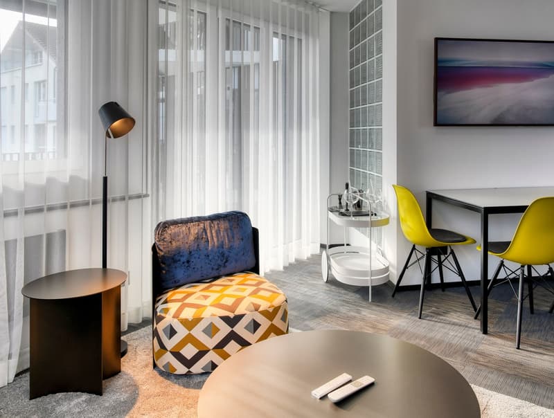 Modern living room with large windows, patterned armchair, side table, floor lamp, and a dining area with a table and yellow chairs