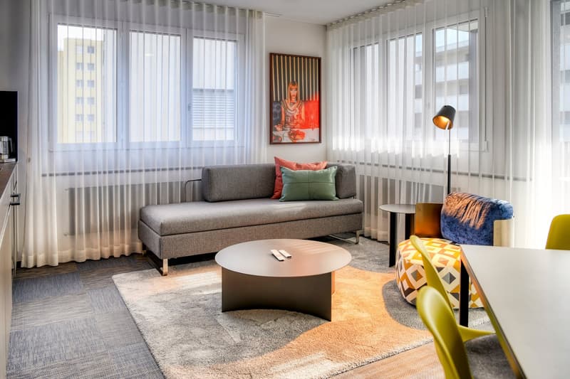 Spacious living room with large windows, gray sofa, round coffee table, colorful artwork on the wall, and a floor lamp