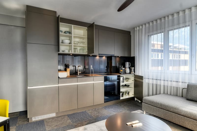 Modern kitchen with gray cabinets, stainless steel appliances, open shelving, and large windows providing natural light