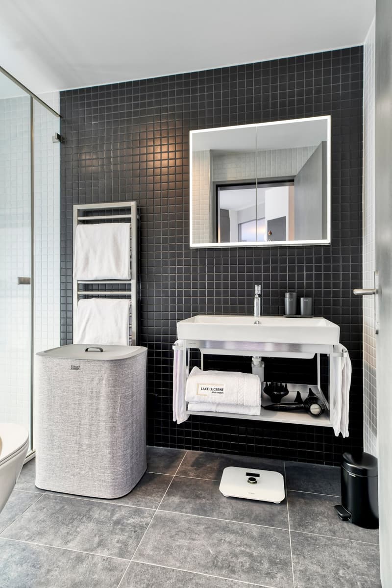 Modern bathroom with black tile walls, white vanity with sink, mirror, towel rack, and a gray laundry hamper.