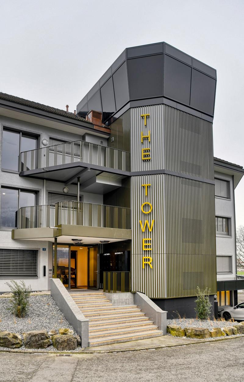 A modern multi-story building with a distinctive black and yellow facade. The building has balconies on the upper floors and a prominent entrance with the word 'Tower' displayed in large yellow letters.