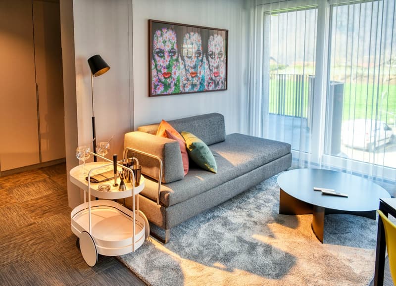 Modern living room with gray sofa, colorful artwork, coffee table, and a bar cart with glasses and bottles