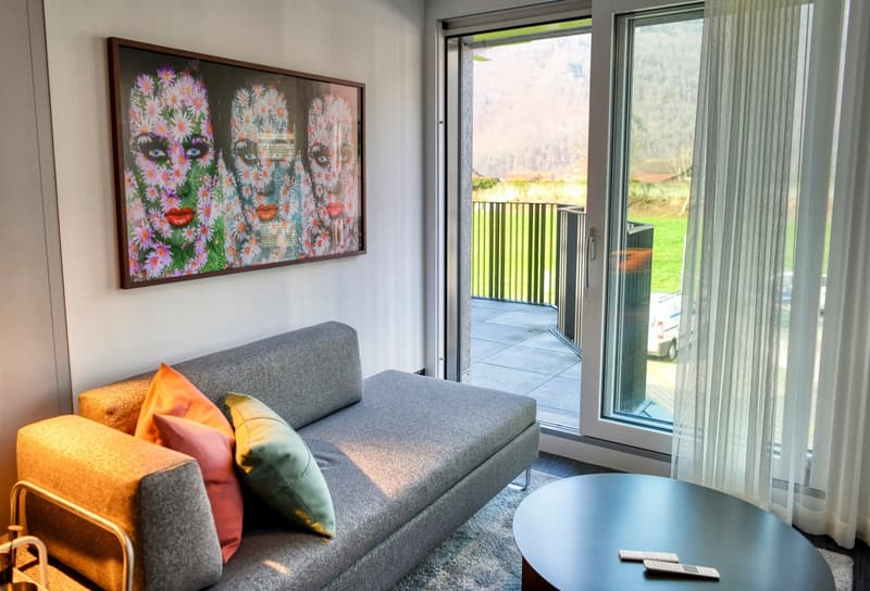 Spacious living room with large windows overlooking a balcony or terrace, gray sofa with colorful throw pillows, framed artwork on the wall depicting floral faces, and a coffee table in the center of the room.