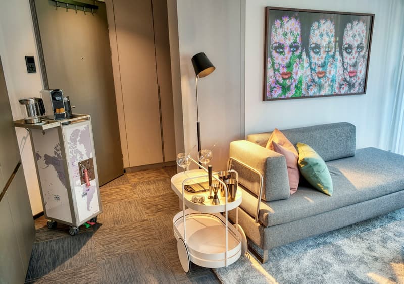 Modern living room with gray sofa, colorful artwork, coffee table with bar accessories, and a kitchenette area with a refrigerator and coffee machine