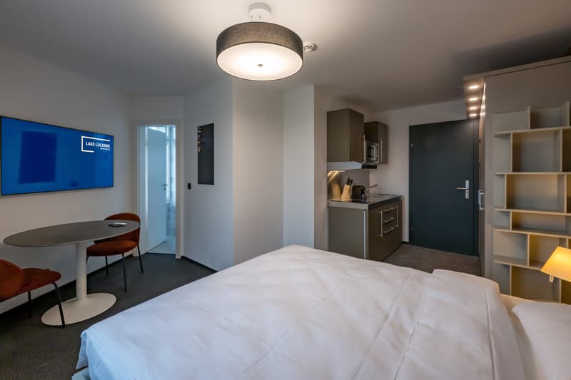 Spacious hotel room with a large bed, a dining table, a kitchenette with a dishwasher, and a large flat-screen TV on the wall.