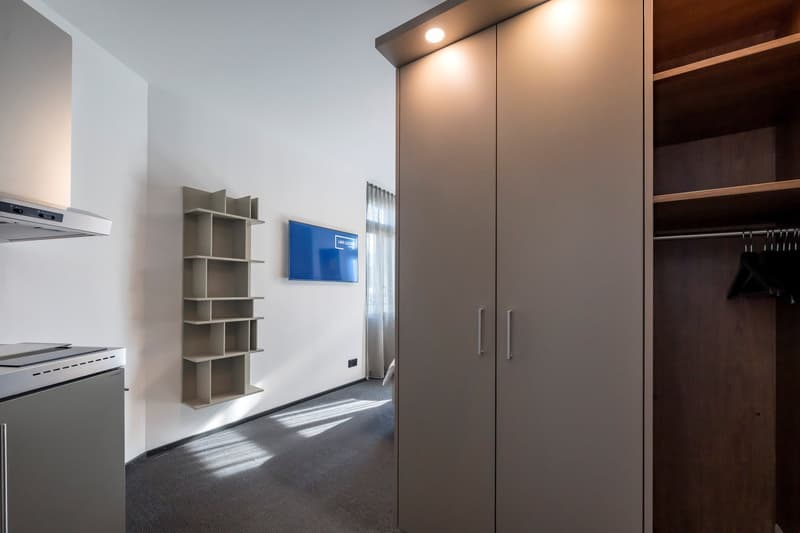 Modern and minimalist interior design with a white wall, floor, and ceiling, shelving, a TV mounted on the wall, and a closet with interior lighting.