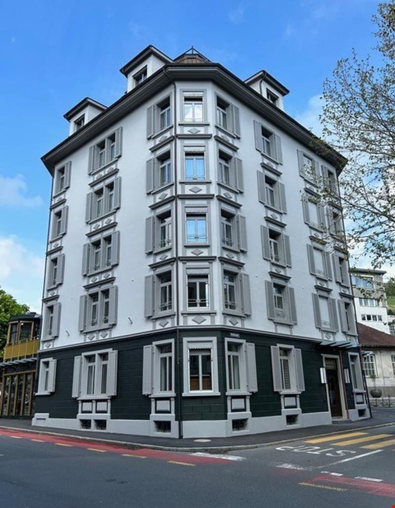 5-story gray and black apartment building with ornate architectural details, including bay windows and decorative moldings. The building has a corner entrance and is situated on a street with a road marking visible.