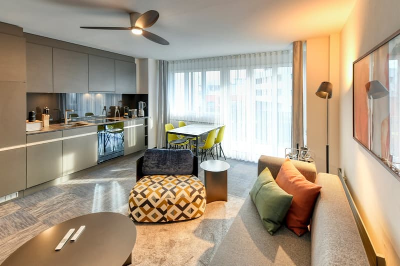 Open-concept living room and kitchen, large windows, modern gray cabinets, stainless steel appliances including a dishwasher, dining table with yellow chairs, patterned ottoman, and couch with colorful pillows