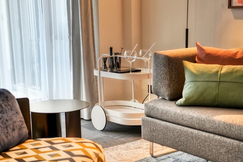 Cozy living room with large windows, gray sofa, colorful throw pillows, coffee table, and a mobile bar cart with wine glasses and bottles