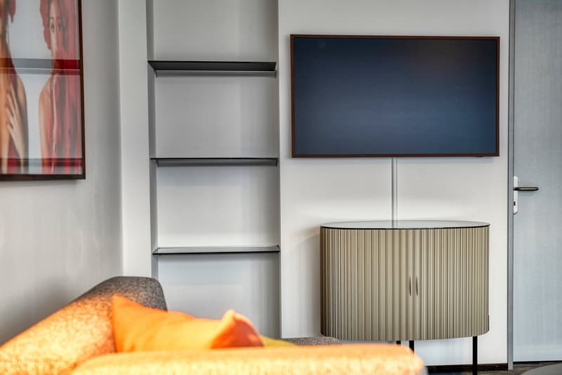 Modern living room with large TV, shelving unit, and a lamp on a side table. The room also features a couch with orange and gray pillows.