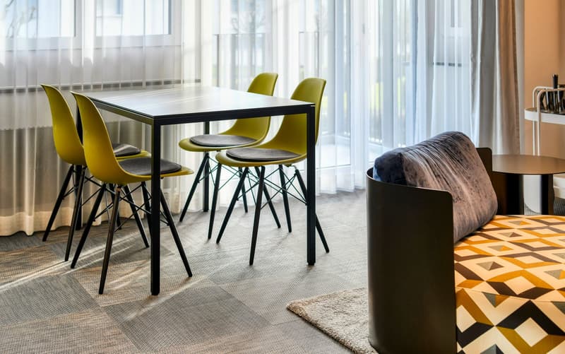 Modern dining area with a black metal table and yellow chairs, large windows providing natural light, patterned rug on the floor, and a cozy sofa in the background.