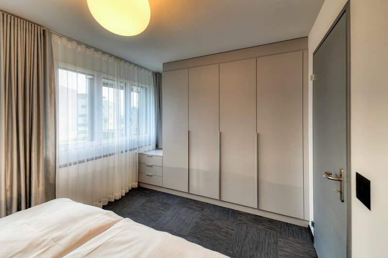 Spacious bedroom with large windows, beige walls, built-in wardrobe, and dark gray carpeted floor.