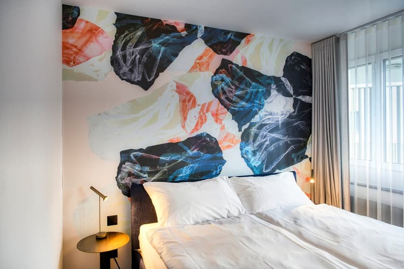 The image shows a modern bedroom with a large, colorful abstract mural covering the wall behind the bed. The mural features vibrant shades of orange, blue, and black, creating a dynamic and visually striking design. The room has a minimalist style, with a