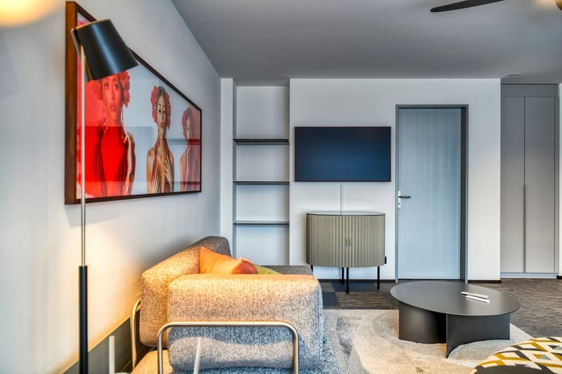 Modern living room with gray sofa, colorful artwork, TV stand, and coffee table. The room has a minimalist and contemporary design with a fireplace and potential access to a balcony or terrace.