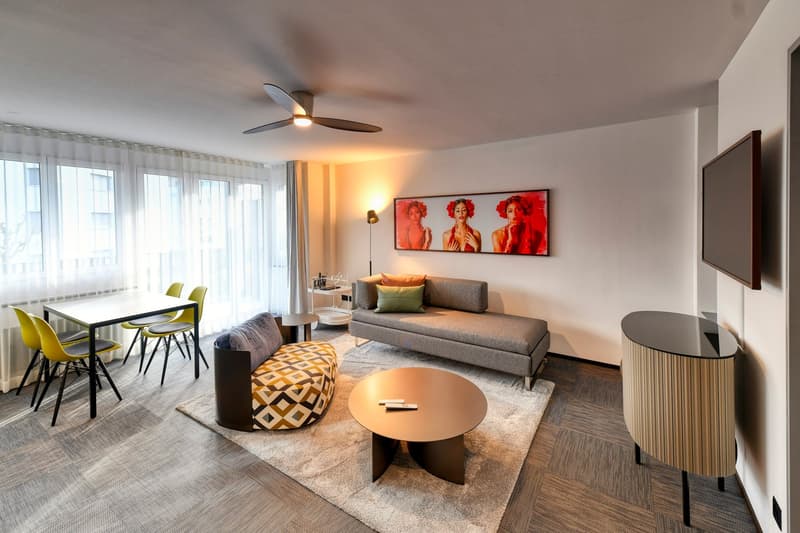 Spacious living room with large windows, gray sofa, patterned pouf, round coffee table, dining table with yellow chairs, ceiling fan, and a large wall art piece featuring three women with red hair.