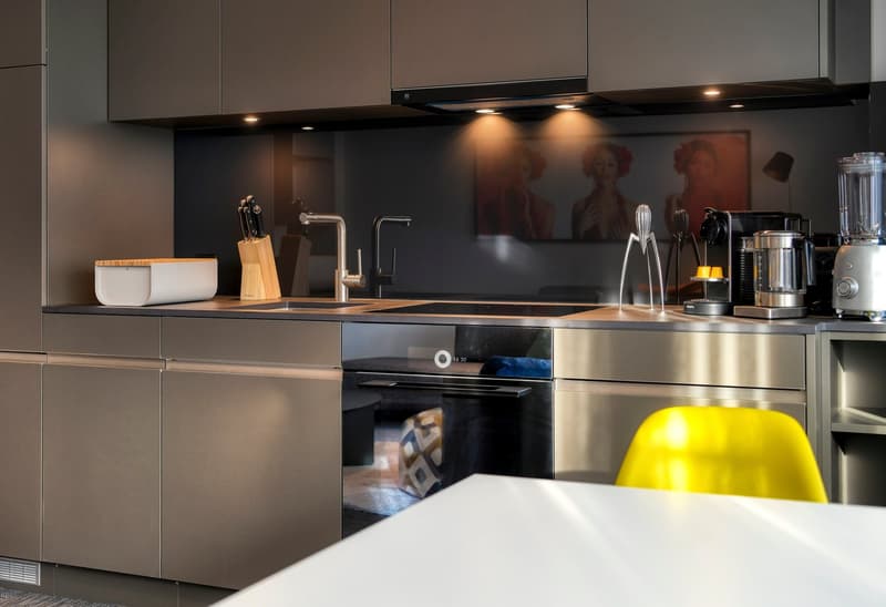 Modern kitchen with dark cabinets, wooden countertops, built-in appliances including a dishwasher, and decorative artwork on the backsplash. The kitchen also features a white table and a large yellow object, possibly a lemon, on the counter.