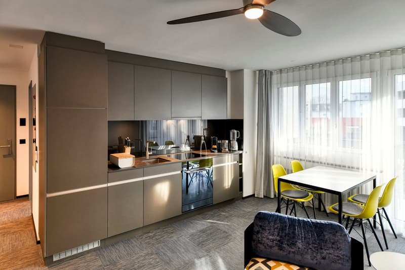 Modern kitchen with gray cabinets, stainless steel appliances including a dishwasher, large windows, and a dining area with a white table and yellow chairs.