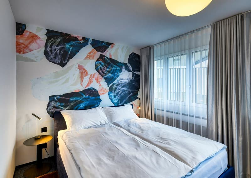 Spacious bedroom with a large abstract mural on the wall, featuring vibrant colors and patterns. The room has a comfortable-looking bed with white bedding, a large window with curtains, and a bedside table with a lamp.