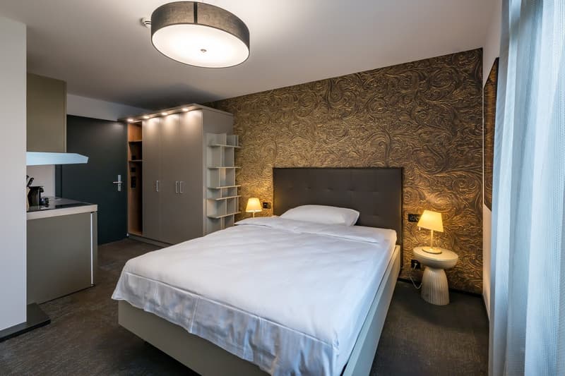 Spacious bedroom with a large bed, a patterned accent wall, built-in shelving, and a kitchenette area with a dishwasher.
