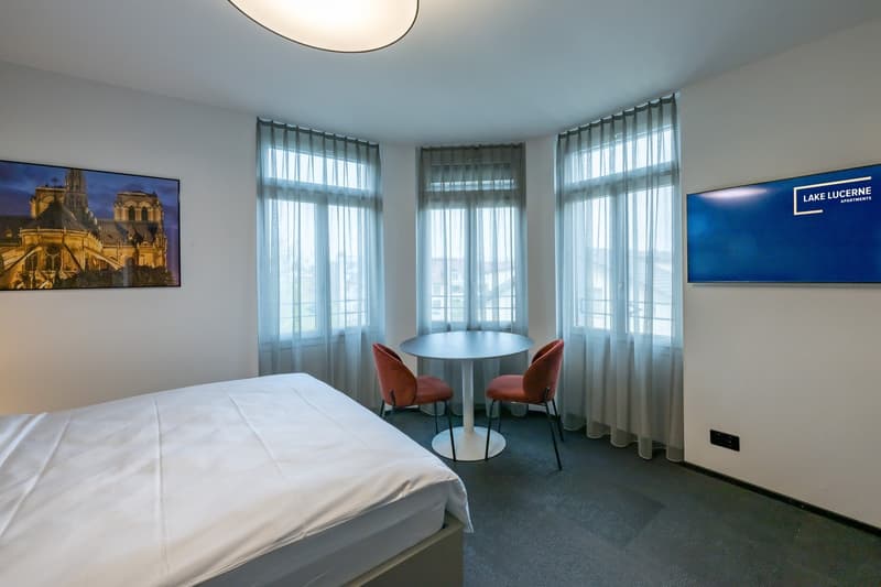Spacious bedroom with large windows providing a view, a round table with two chairs, and artwork on the walls depicting a scenic landscape.