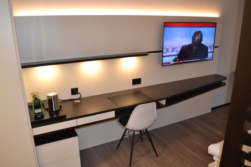 The image shows a modern home office setup with a large desk, a TV mounted on the wall, and various decor items like a wine bottle and a canister. The room has a minimalist and sleek design with white walls, dark wood flooring, and recessed lighting.