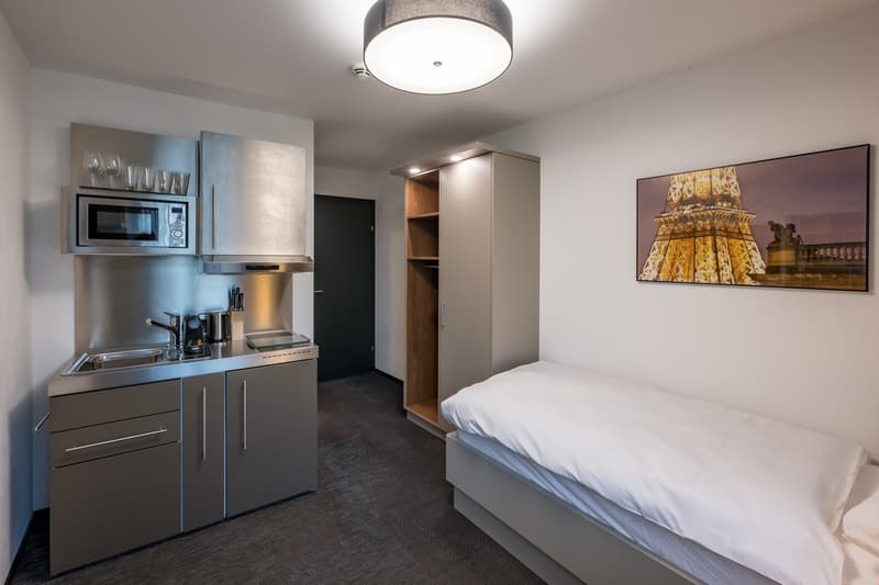 compact room, bed, wardrobe, small kitchenette, silver cabinets, sink, microwave, picture of Eiffel Tower on the wall