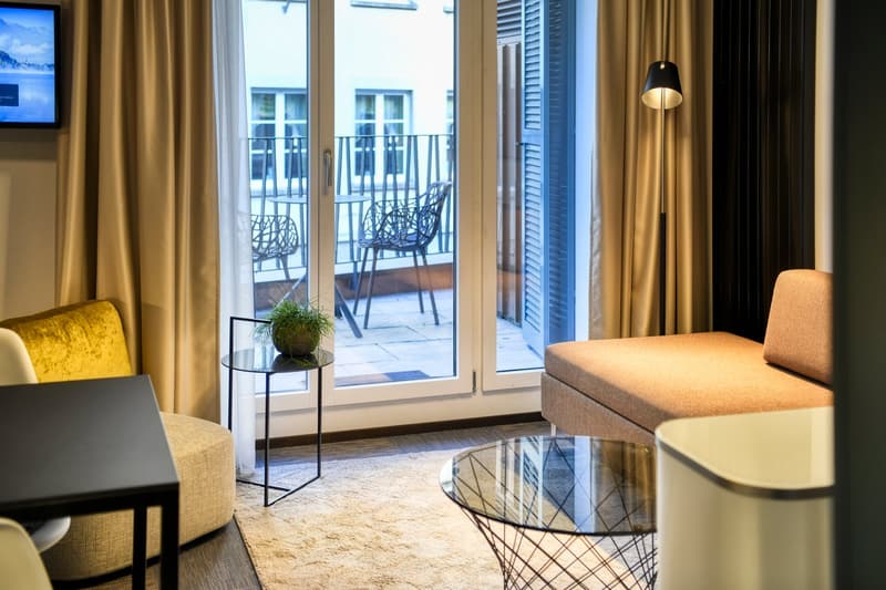 Spacious living room with large windows overlooking a balcony, comfortable yellow armchair, glass coffee table, and a TV mounted on the wall.