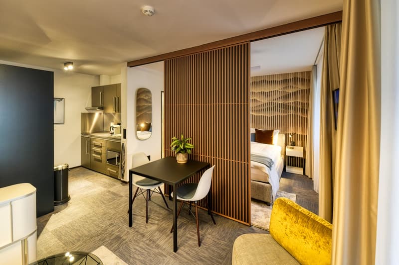 Open-concept studio apartment with kitchenette, dining area, and bedroom area separated by a wooden slatted partition. The space features hardwood-style flooring, a yellow velvet sofa, and a small dining table with two chairs. The kitchenette includes sta