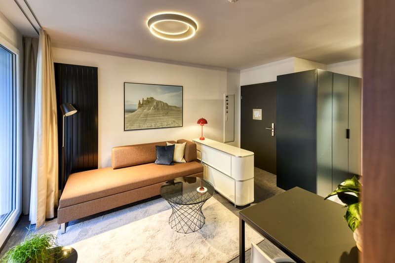 Modern living room with large sofa, coffee table, dresser, and framed artwork on the wall. The room has a minimalist and contemporary design with a circular lighting fixture on the ceiling. There is a large window that suggests the presence of a balcony o