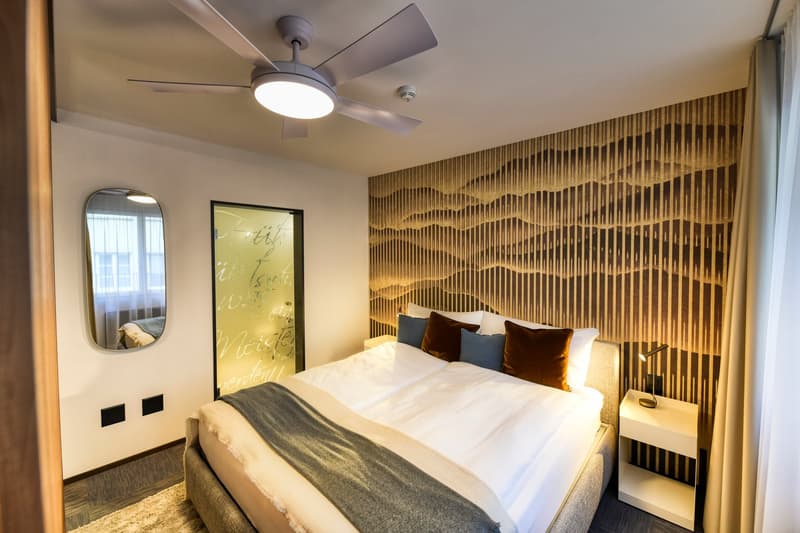 Modern bedroom with a textured accent wall, a large window, a ceiling fan, a bed with colorful pillows, and a framed artwork on the wall.