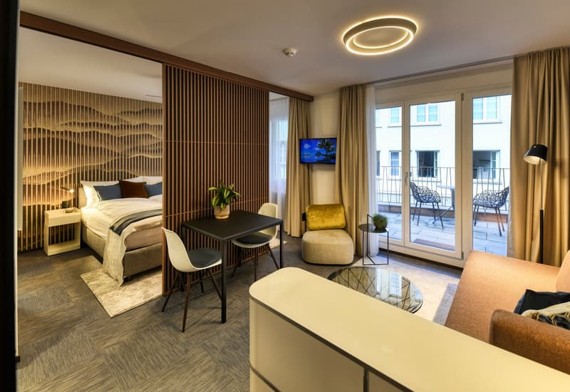 Spacious hotel room with a modern and cozy design. The room features a large bed, a seating area with a table and chairs, and a balcony with outdoor furniture. The room has wooden paneling on the walls, creating a warm and inviting atmosphere. There is al