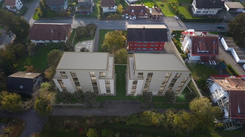 Two multi-story apartment buildings with white facades, surrounded by trees and greenery, with a road and other residential buildings visible in the background.