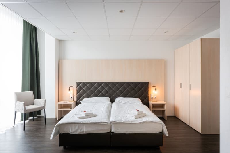Spacious bedroom with a large bed, quilted headboard, and side tables. The room has a modern and minimalist design with white walls, a suspended ceiling, and wooden flooring. There is a window with green curtains, suggesting the presence of a balcony or t