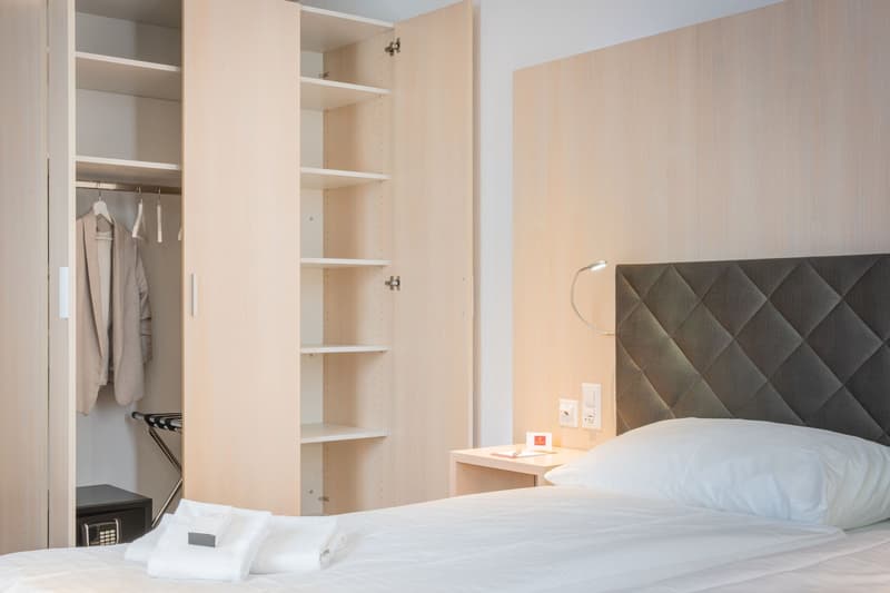 Bedroom with built-in closet, shelves, and a quilted headboard. There is a small nightstand with a lamp and a safe. The room also has a washing machine and a dishwasher.