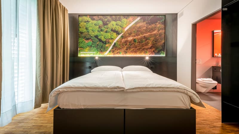 Large bed with white bedding, wooden floor, curtains, and a large screen displaying a scenic landscape view