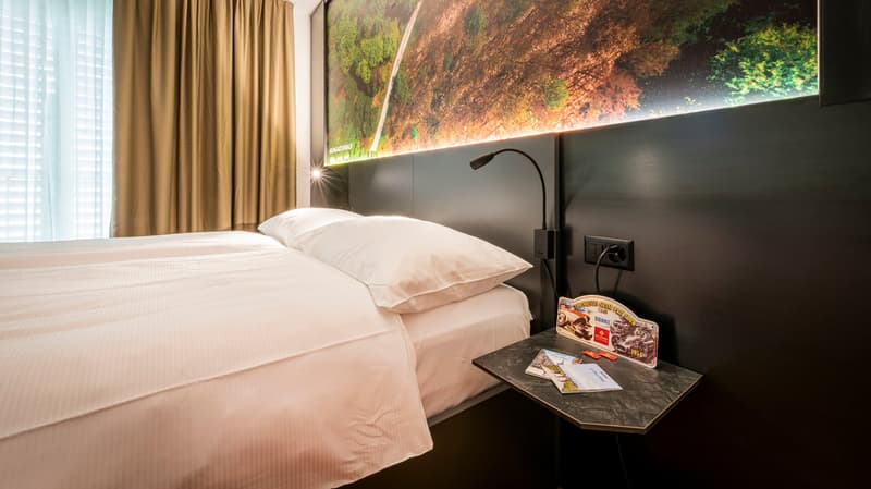 Spacious bedroom with large window displaying a scenic landscape, white bedding, dark headboard, bedside table with lamp, and decorative item on the table