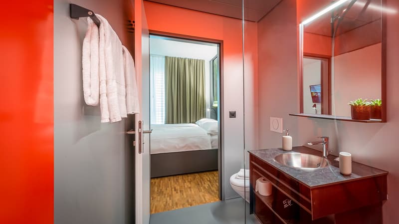 Spacious bathroom with red walls, wooden floor, vanity with marble countertop, sink, and mirror. Bedroom visible through the open doorway.