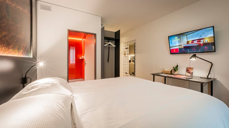 A bedroom with a large bed, a desk with a lamp, a TV mounted on the wall, a wardrobe, and a red door.